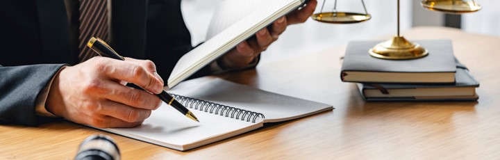 Person in Anzug sitzt an einem Tisch, auf dem ein Heft liegt, in das die Person etwas aufschreiben möchte. Neben ihm liegen zwei Bücher aufeinander gestapelt, auf denen eine Waage steht, die nur teilweise abgebildet ist.
