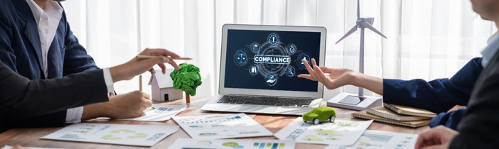 Blick auf einen Tisch, auf dem mehrere Blätter liegen und ein Laptop steht. Auf dem Bildschirm des Laptops steht das Wort Compliance steht. An dem Tisch sitzen mehrere Personen, die zu diskutieren scheinen. 