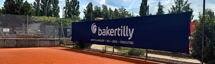 Blick vom Tennisplatz auf das Baker Tilly Logo, das vor grünen Bäumen am Zaun hängt. 