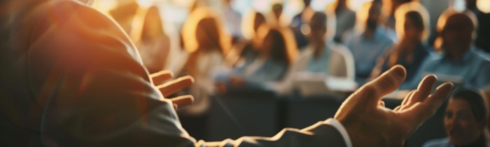 im vordergrund sieht man zwei ausgebreitete arme. der hintergrund ist gefüllt mit leuten die dem mann mit den ausgebreiteten armen zuhören