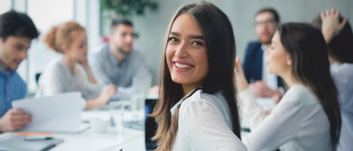 Mehrere Kolleginnen und Kollegen sitzen an einem Tisch, auf dem verschiedene Dokumenten liegen und durchgeblättert werden. Eine junge Frau blickt dabei direkt auf den Betrachter.