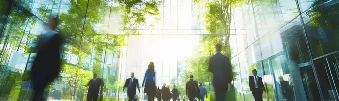 In einer modernen, lichtdurchfluteten Glasarchitektur bewegen sich mehrere Menschen in Businesskleidung durch einen offenen Innenraum. Die Personen sind dabei teils unscharf dargestellt. Große Glasflächen spiegeln grüne Bäume und Tageslicht, sodass Innen- und Außenraum miteinander verschmelzen.