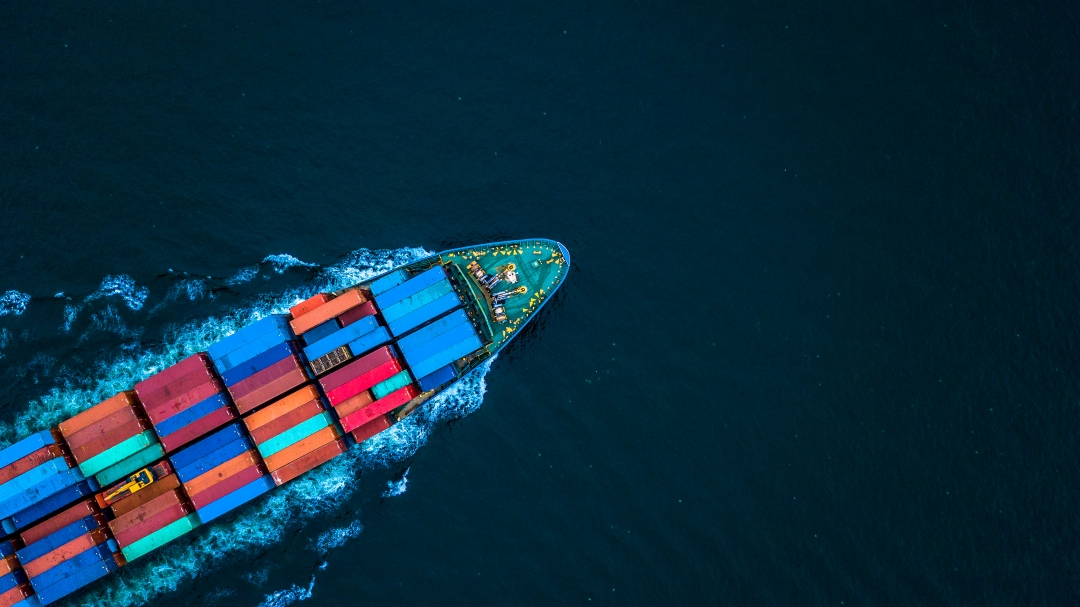 Ein Containerschiff aus der Vogelperspektive. Es fährt durch das türkisfarbene Meer.