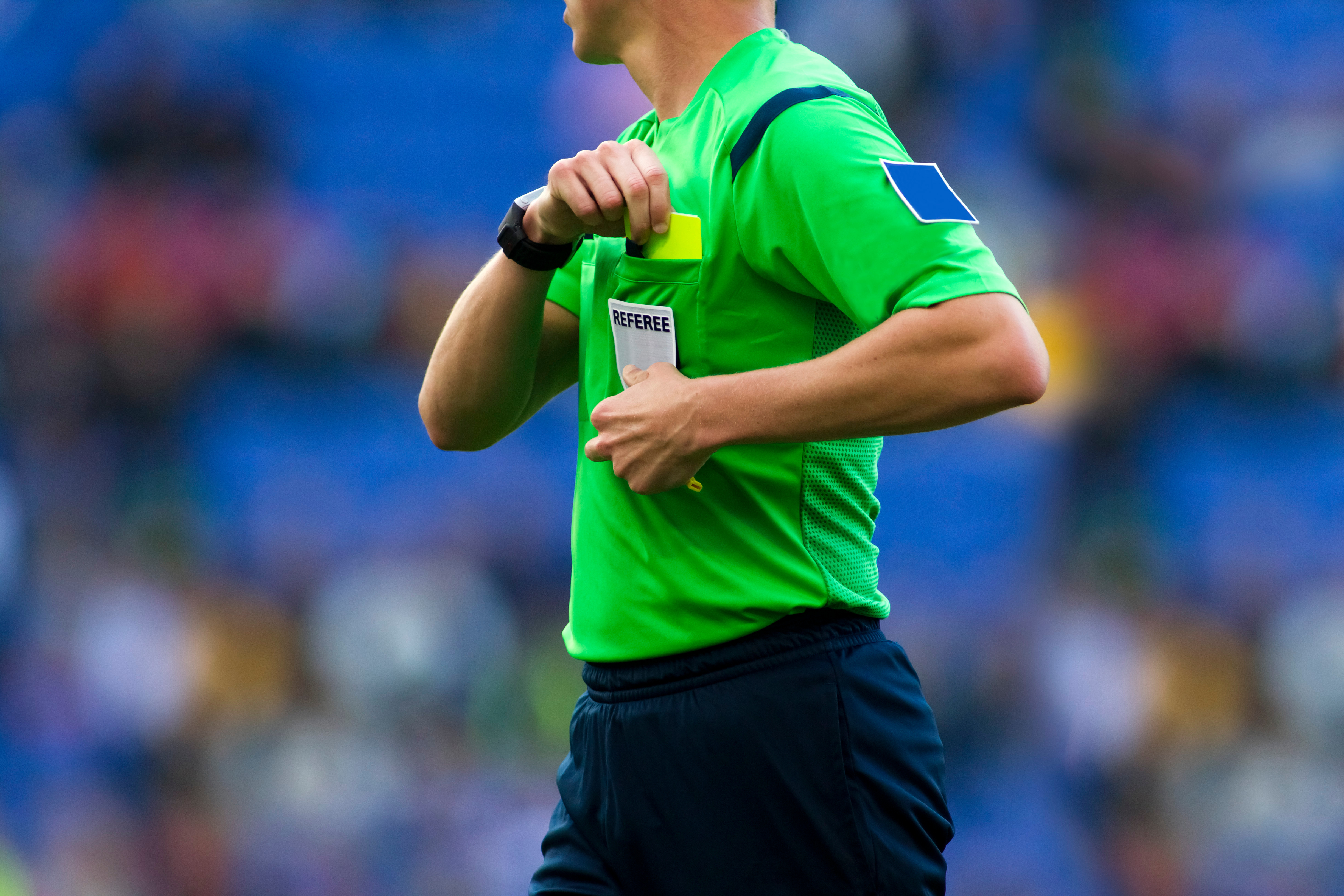 Foto: Ein Fußball-Schiedsrichter im hellgrünen Referee-Trikot greift zur linken Brusttasche. Darin befindet sich die Gelbe Karte.