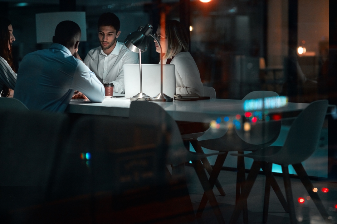 Foto: Eine Gruppe von Mitarbeitern eines Start-ups sitzt in einem dunklen Büro an einem Konferenztisch, der von einer einzigen Schreibtischlampe erleuchtet ist.