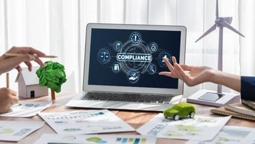 Blick auf einen Tisch, auf dem mehrere Blätter liegen und ein Laptop steht. Auf dem Bildschirm des Laptops steht das Wort Compliance steht. An dem Tisch sitzen mehrere Personen, die zu diskutieren scheinen. 