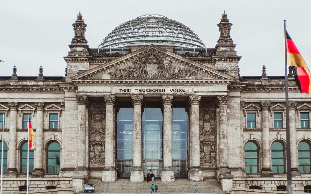 Foto: Der Reichstag in Berlin, vor grau bewölktem Himmel. Davor weht eine Deutschland-Fahne.