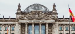 Foto: Der Reichstag in Berlin, vor grau bewölktem Himmel. Davor weht eine Deutschland-Fahne.