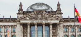 Foto: Der Reichstag in Berlin, vor grau bewölktem Himmel. Davor weht eine Deutschland-Fahne.