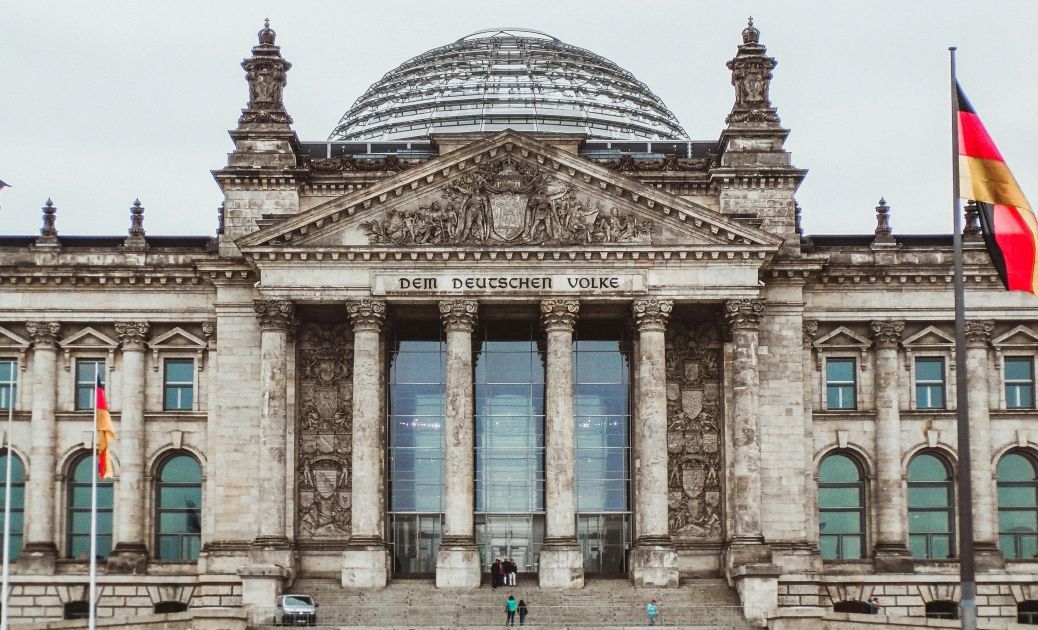 Foto: Der Reichstag in Berlin, vor grau bewölktem Himmel. Davor weht eine Deutschland-Fahne.