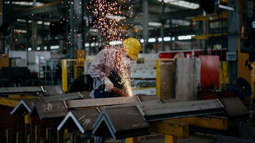 Foto: Ein Arbeiter macht sich in einer Produktionshalle mit einer Flex an einem Metallteil zu schaffen. Funken fliegen.