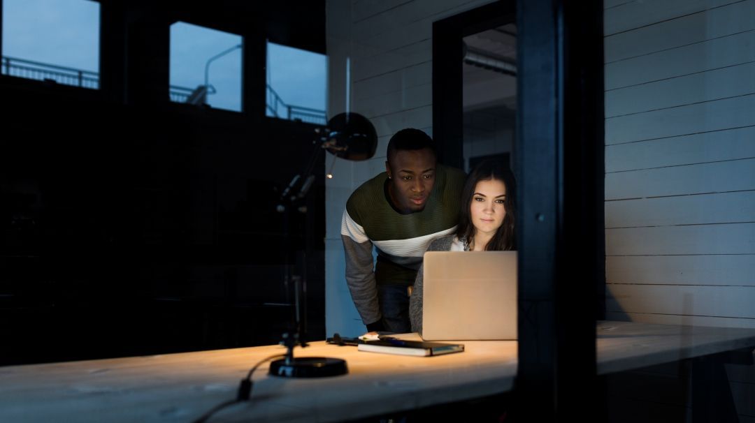 Foto: Zwei Kollegen sitzen in einem dunklen Büro vor einem Laptop. Eine einzelne Schreibtischlampe brennt.