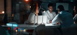 Foto: Eine Gruppe von Mitarbeitern eines Start-ups sitzt in einem dunklen Büro an einem Konferenztisch, der von einer einzigen Schreibtischlampe erleuchtet ist.
