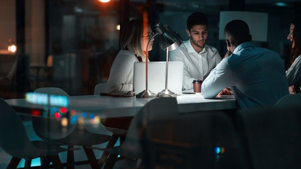 Foto: Eine Gruppe von Mitarbeitern eines Start-ups sitzt in einem dunklen Büro an einem Konferenztisch, der von einer einzigen Schreibtischlampe erleuchtet ist.