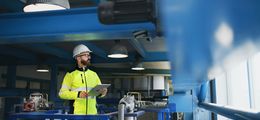 Foto: Ein Mann mit Schutzhelm und Hi-Vis Schutzkleidung steht in einer Fabrik. Er hat ein Klemmbrett in der Hand.