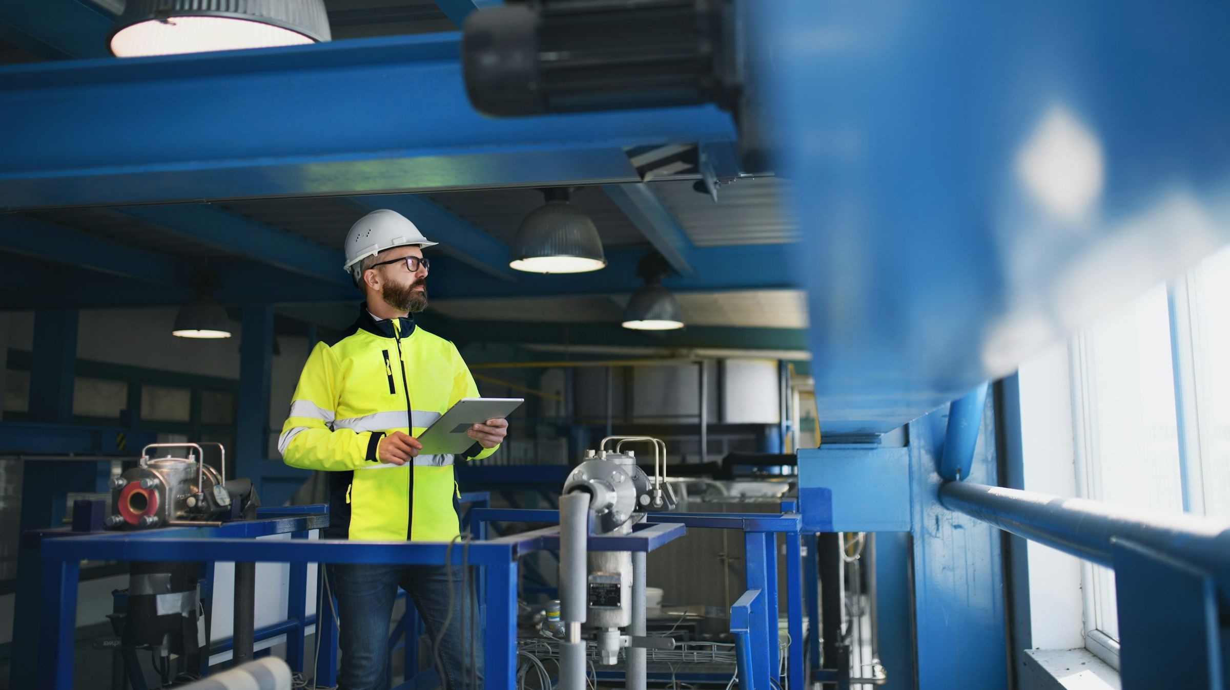 Foto: Ein Mann mit Schutzhelm und Hi-Vis Schutzkleidung steht in einer Fabrik. Er hat ein Klemmbrett in der Hand.