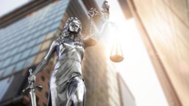 Links sieht man Justitia vor einem Bürogebäude, die in die Flagge der EU übergeht.