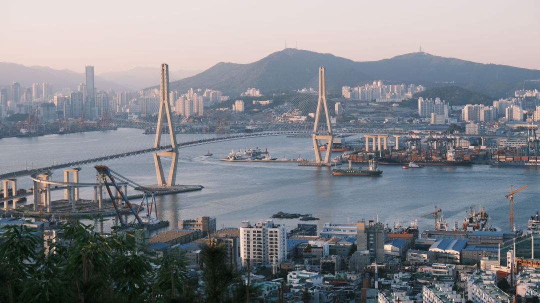 Foto der südkoreanischen Hafenstadt Busan. Es fällt ein mildes Licht auf die Brücke und Skyline. Im Hintergrund die Berge.