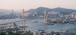 Foto der südkoreanischen Hafenstadt Busan. Es fällt ein mildes Licht auf die Brücke und Skyline. Im Hintergrund die Berge.