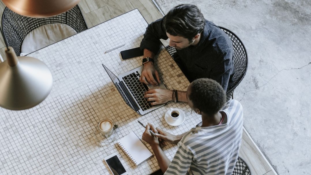 Foto: Zwei Kollegen sitzen gemeinsam an einem Tisch. Ein Laptop steht zwischen ihnen.