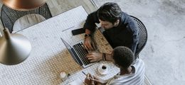Foto: Zwei Kollegen sitzen gemeinsam an einem Tisch. Ein Laptop steht zwischen ihnen.