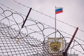 Foto: Stacheldrahtzaun. Im Hintergrund weht die russische Flagge auf einem Regierungsgebäude.