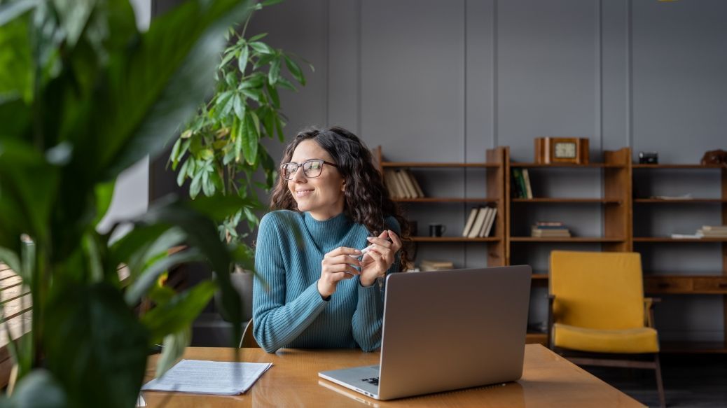 Foto: Eine Frau sitzt lächelnd an einem Arbeitstisch und schau aus dem Fenster. Vor ihr steht auf aufgeklappter Laptop.