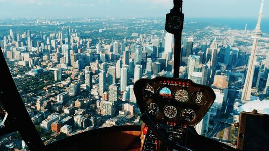 Die Aufnahme zeigt den Blick aus dem Cockpit eines Hubschraubers auf eine dichte Skyline mit zahlreichen hohen Gebäuden, die sich bis zum Horizont ausbreiten. Rechts im Bild ragt ein sehr schlanker, nadelförmiger Turm deutlich in den Himmel. Weiter hinten ist ein großer Wasserbereich sichtbar. Im Vordergrund sind die Cockpit-Instrumente deutlich zu sehen – verschiedene Rundanzeigen, Schalter und Hebel.