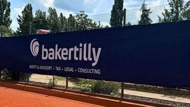 Blick vom Tennisplatz auf das Baker Tilly Logo, das vor grünen Bäumen am Zaun hängt. 