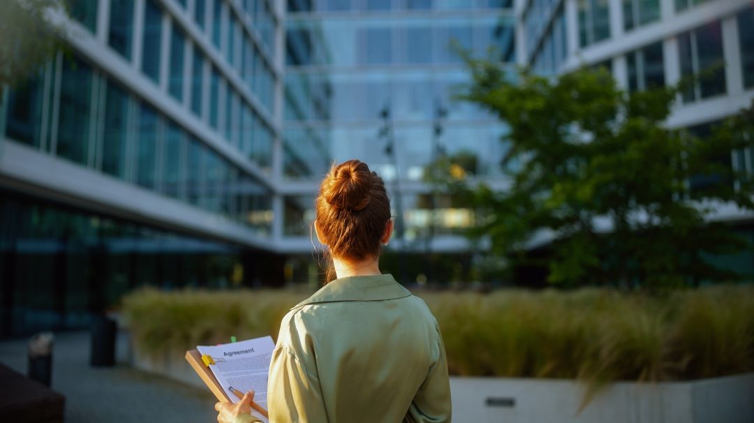 Eine Frau steht vor einem Bürogebäude. Die hält Vertragsunterlagen in der Hand.