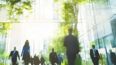 In einer modernen, lichtdurchfluteten Glasarchitektur bewegen sich mehrere Menschen in Businesskleidung durch einen offenen Innenraum. Die Personen sind dabei teils unscharf dargestellt. Große Glasflächen spiegeln grüne Bäume und Tageslicht, sodass Innen- und Außenraum miteinander verschmelzen.