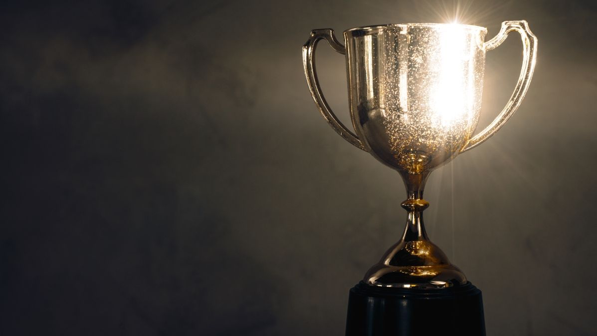 Foto: Ein goldener, glänzender Pokal steht vor einer dunklen Wand.
