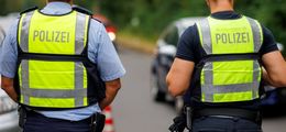 Foto: Zwei Polizisten mit Warnwesten stehen an einer Straße. Es werden Grenzkontrollen durchgeführt. Das ist an Autos und Pylonen im Hintergrund zu erkennen ist.