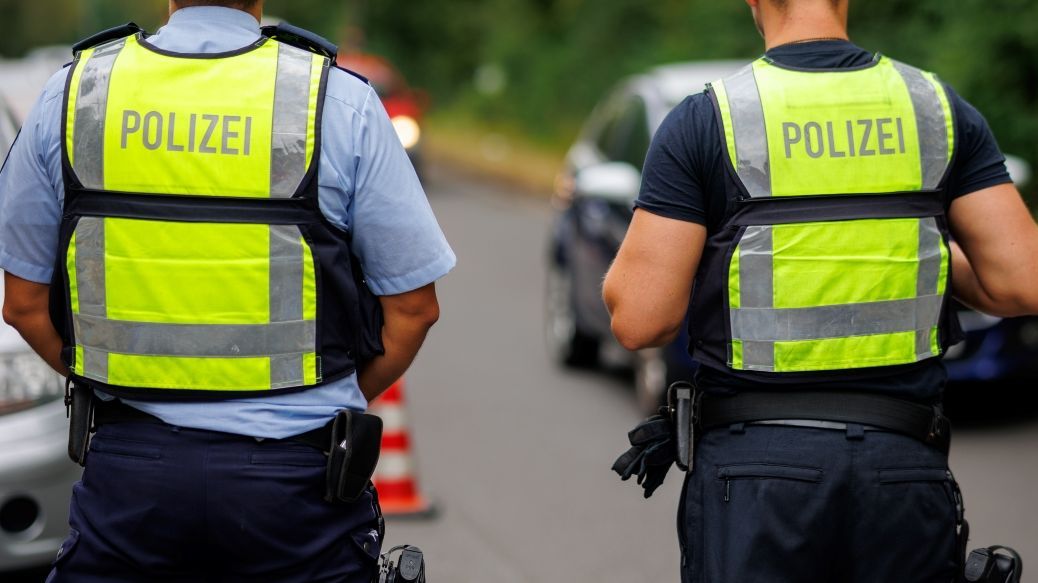 Foto: Zwei Polizisten mit Warnwesten stehen an einer Straße. Es werden Grenzkontrollen durchgeführt. Das ist an Autos und Pylonen im Hintergrund zu erkennen ist.