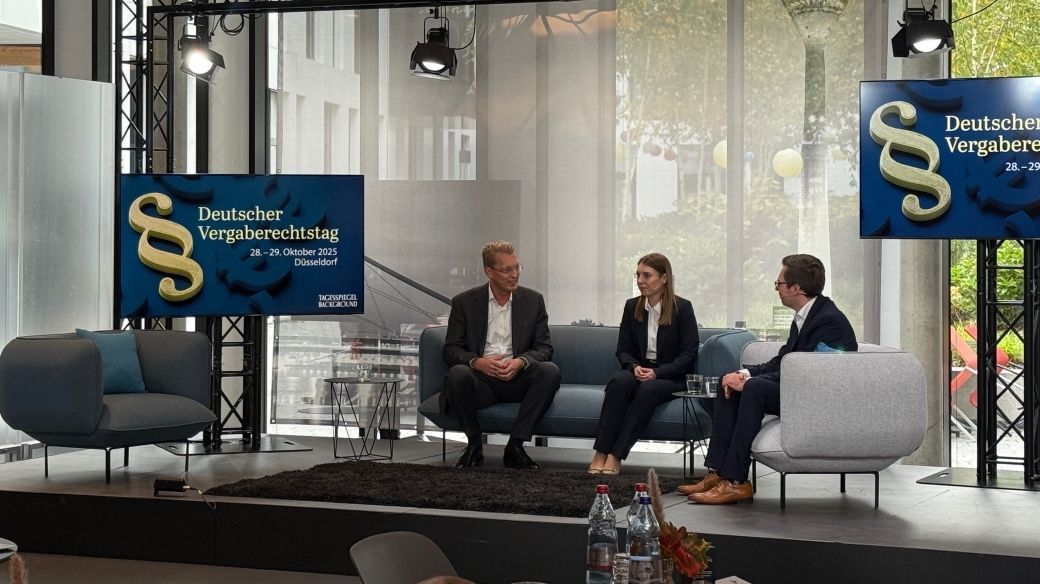 Foto: Dr. Christian Teuber, Elisabeth Bruhn und Dr. Moritz Philipp Koch sitzen auf einem Panel auf einer Bühne. Im Hintergrund ist auf Bildschirmen zu sehen, dass es sich um den Deutschen Vergaberechtstag 2025 von Tagesspiegel Background handelt.
