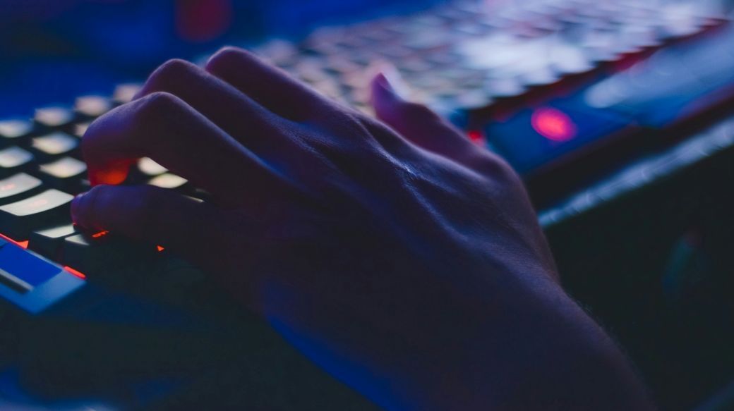Foto: Eine Hand tippt auf einer leuchtenden Tastatur, in einem dunklen Raum.