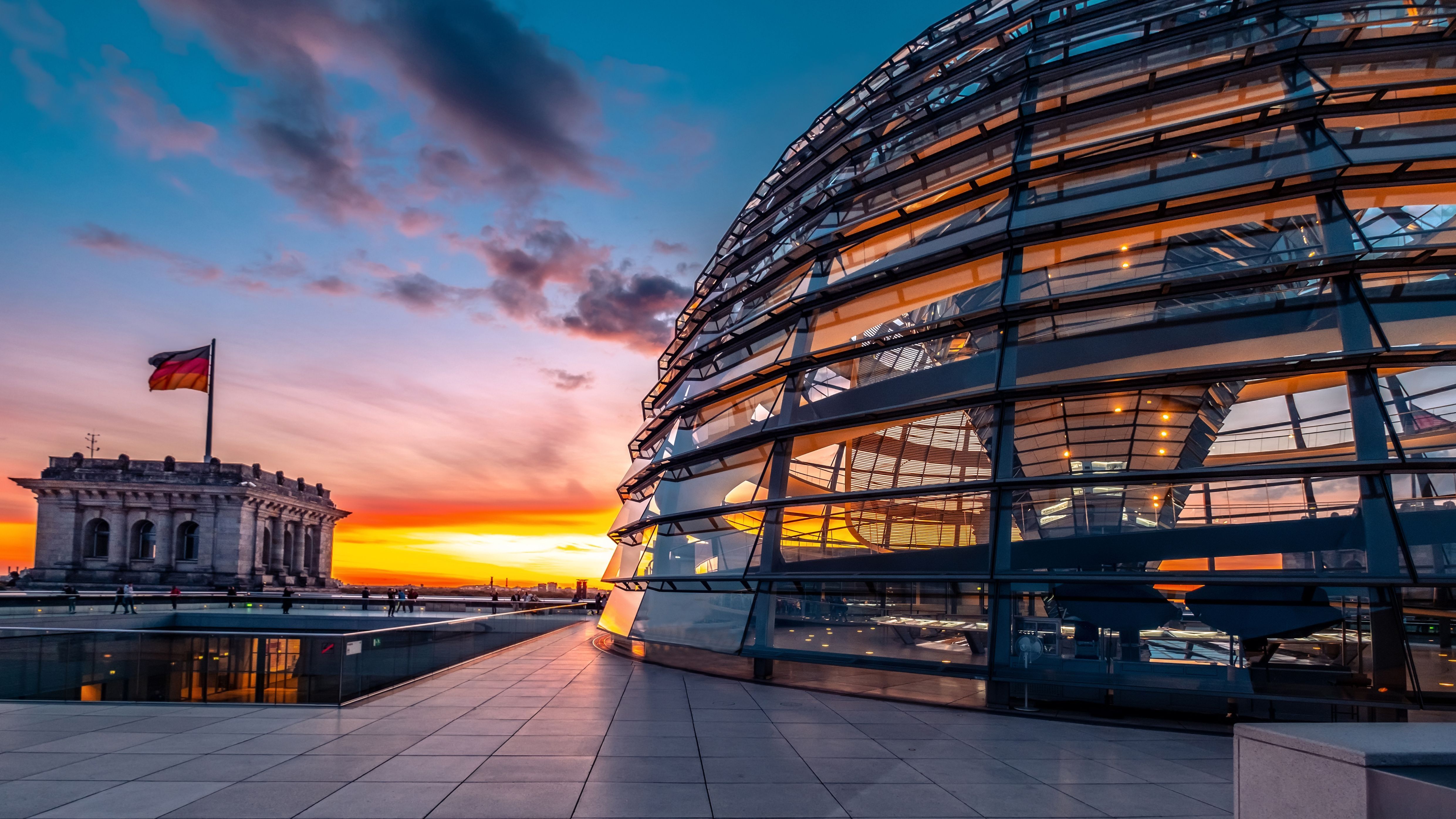 Bild: gläserne Kuppel des Reichstagsgebäudes in Berlin beim Sonnenuntergang und der deutschen Flagge im Vordergrund.