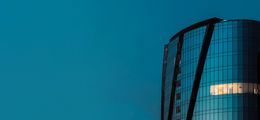Foto: Moderne Bürogebäude mit abgerundeter Fassade in der dunkelblauen Abenddämmerung. Auf einer Etage brennt noch Licht.