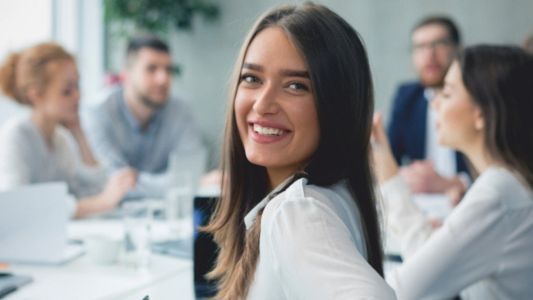 Mehrere Kolleginnen und Kollegen sitzen an einem Tisch, auf dem verschiedene Dokumenten liegen und durchgeblättert werden. Eine junge Frau blickt dabei direkt auf den Betrachter.