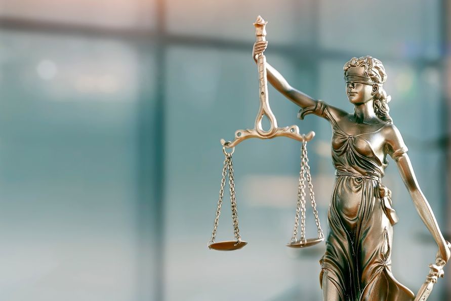 Foto: Eine Messingstatue der Justizia vor unscharfem, hellblauen Hintergrund.